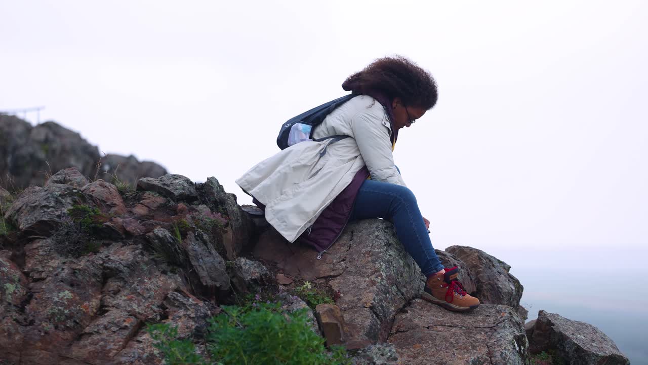 mixed race woman with curly hair is sitting on the ledge of a cliff in Iceland during an overcast day