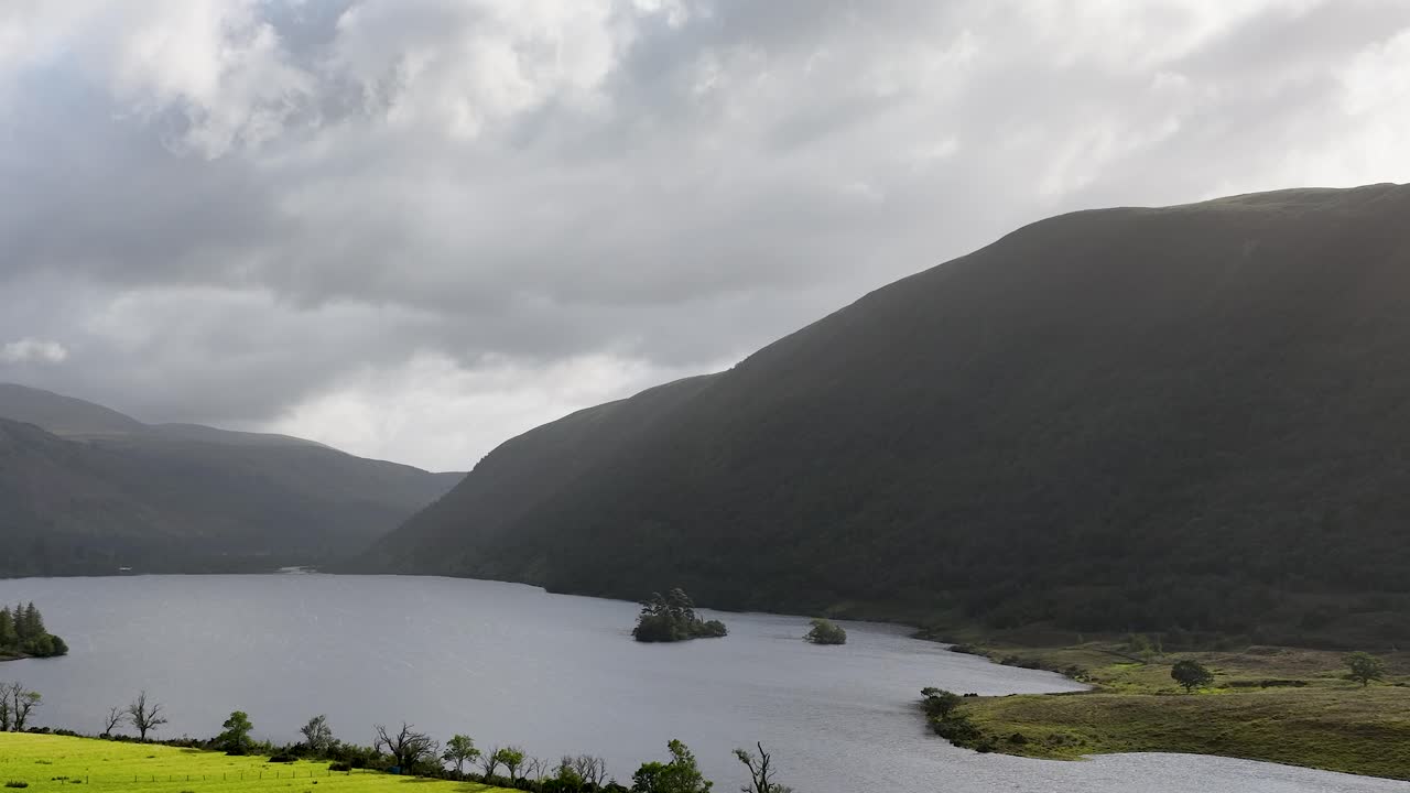 Aerial camera glides over tranquil lake, green meadows, and misty mountains under dramatic clouds