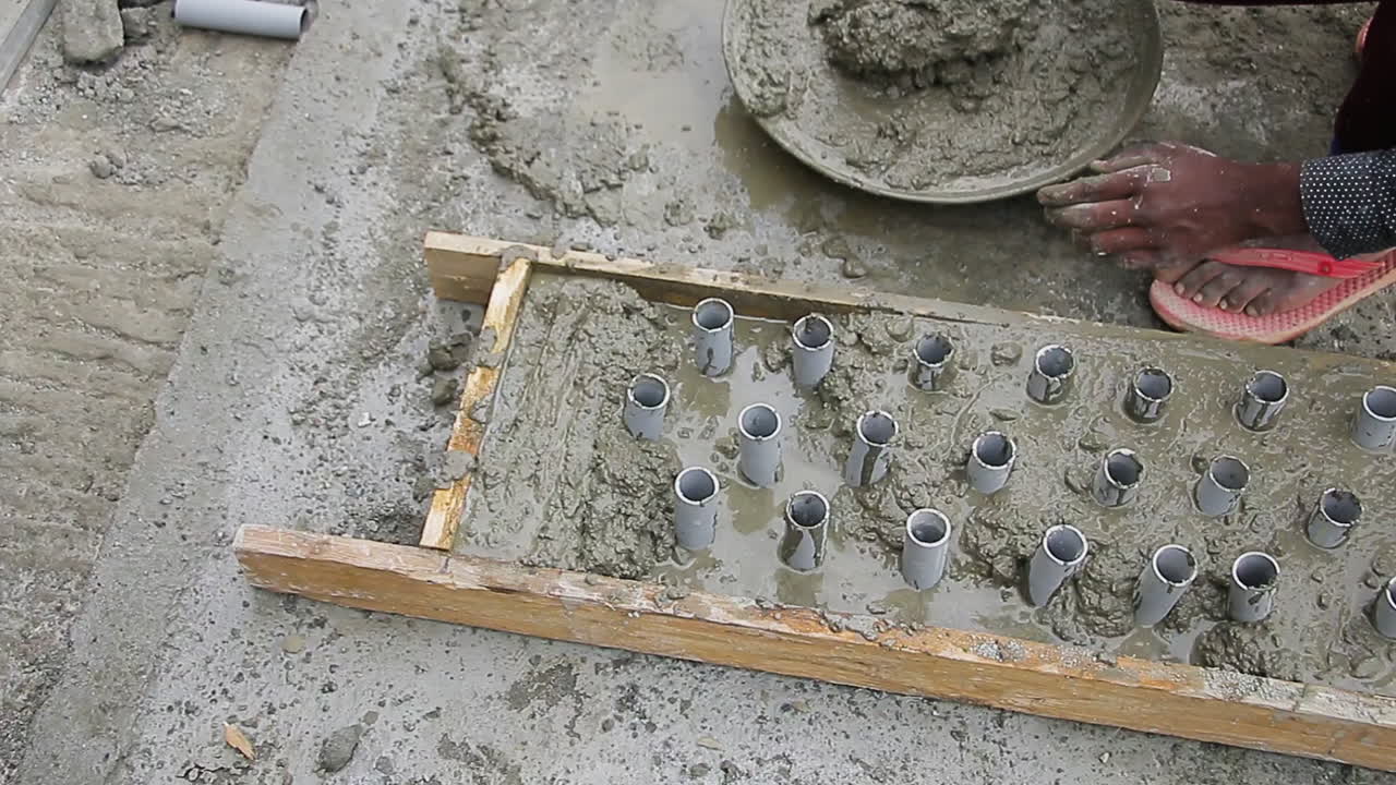 Indian mason worker placing concrete mix around pipes in a wooden form and leveling it for slab construction