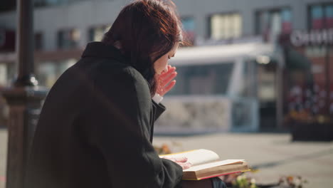 close-up back view of a lady focused on reading her book outdoors as her hair gently sways in the breeze, she flips to the next page while sunlight softly highlights the scene in an urban environment