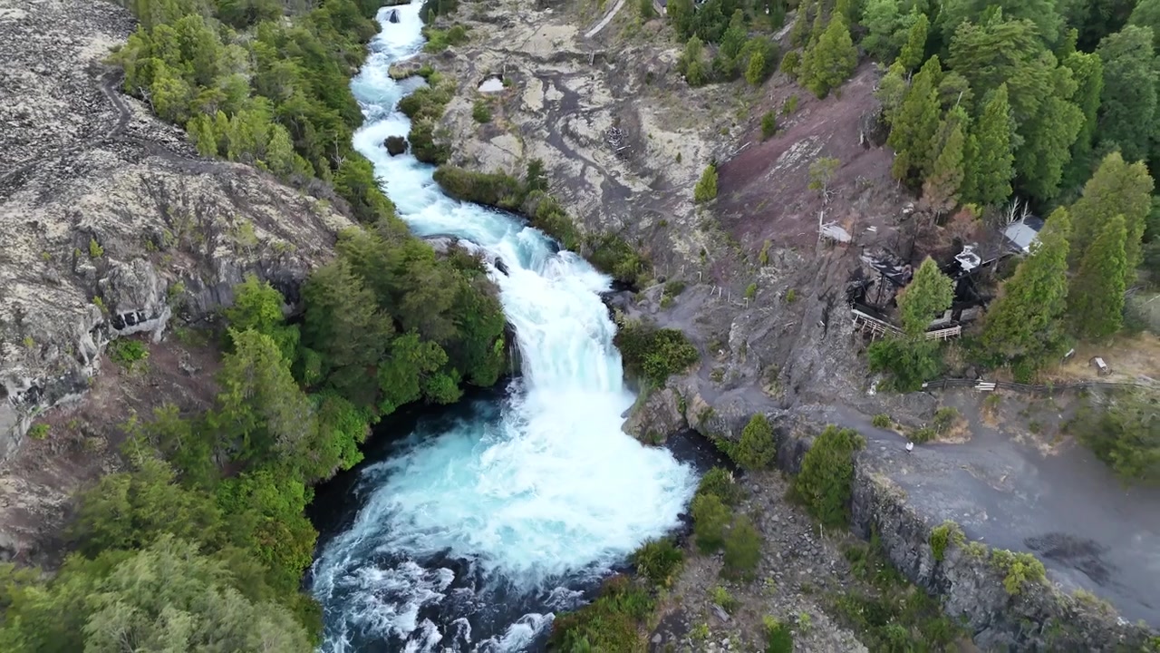 Aerial drone footage pulling back from a dramatic turquoise waterfall cascading through a deep volcanic canyon surrounded by dense conifer forest. Clear blue sky, bright midday sunlight