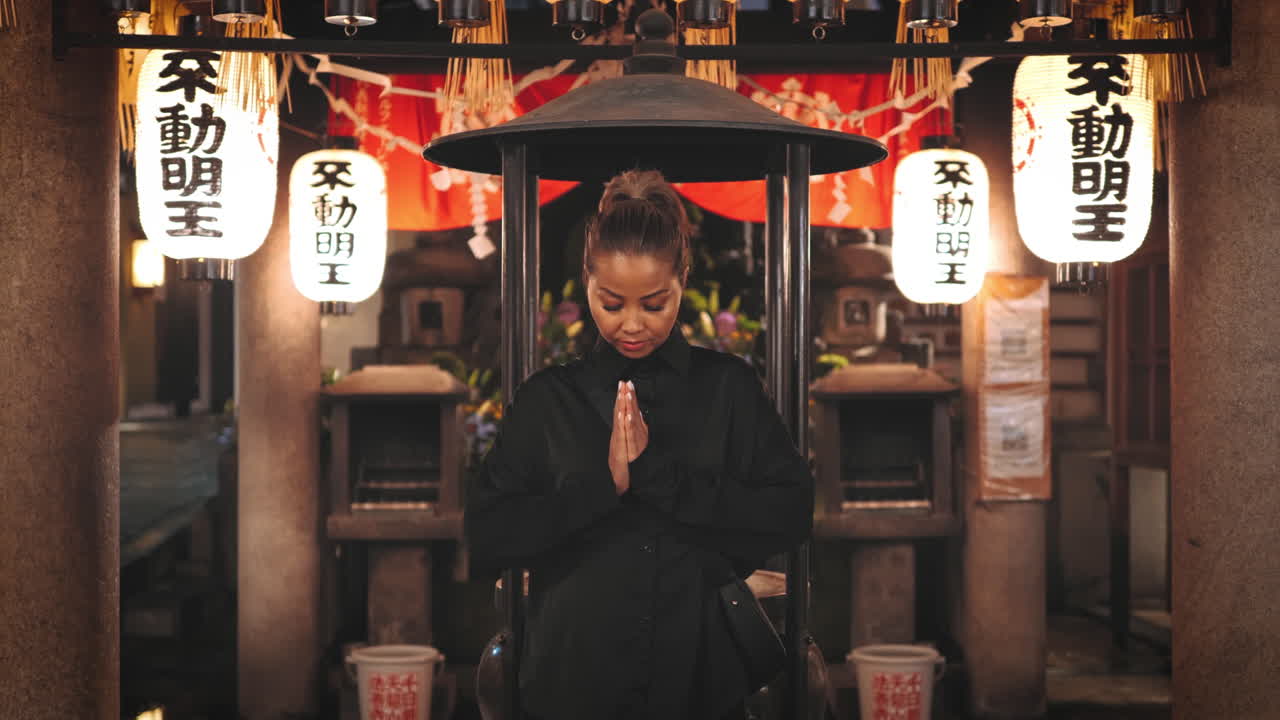 mujer de cabello marrón claro, vestida de negro, de pie con las manos en oración en un santuario japonés