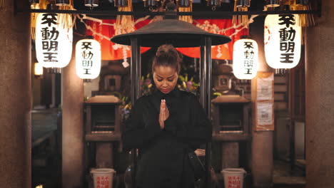 mujer de cabello marrón claro, vestida de negro, de pie con las manos en oración en un santuario japonés