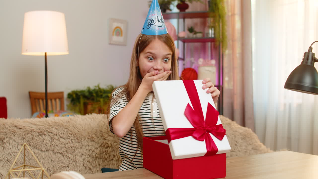 Niña celebra su cumpleaños, abre felizmente un regalo con sorpresa y emoción en el sofá de su casa, junto a la mesa.
