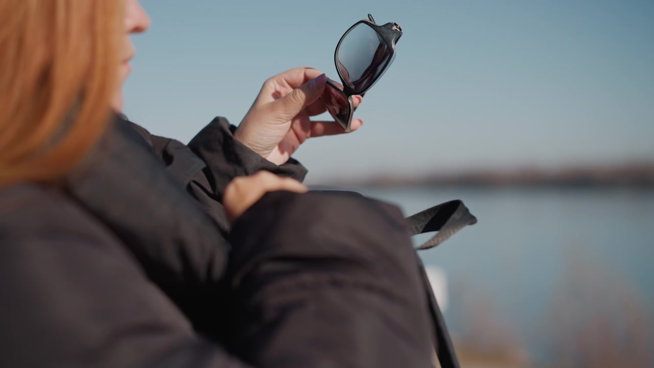 Woman outdoors near water handling black bag and retrieving sunglasses. Woman wears dark clothing with long hair visible from side as woman interacts with her personal items against outdoor