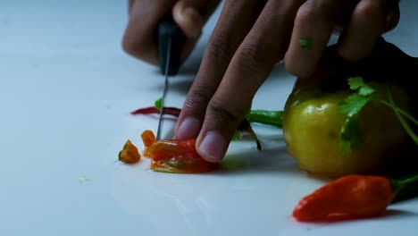 slow motion shot of a man cutting a red bombay chili pepper on top of a white surface, next to other vegetables