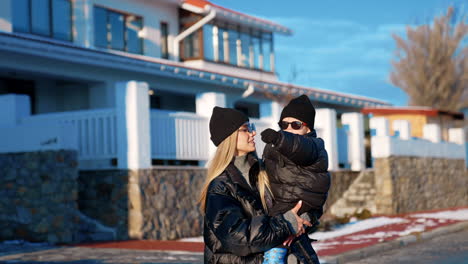 beautiful blonde woman wearing black clothes and sunglasses holds her baby boy in sunglasses too. mother waves her child standing near a big house.