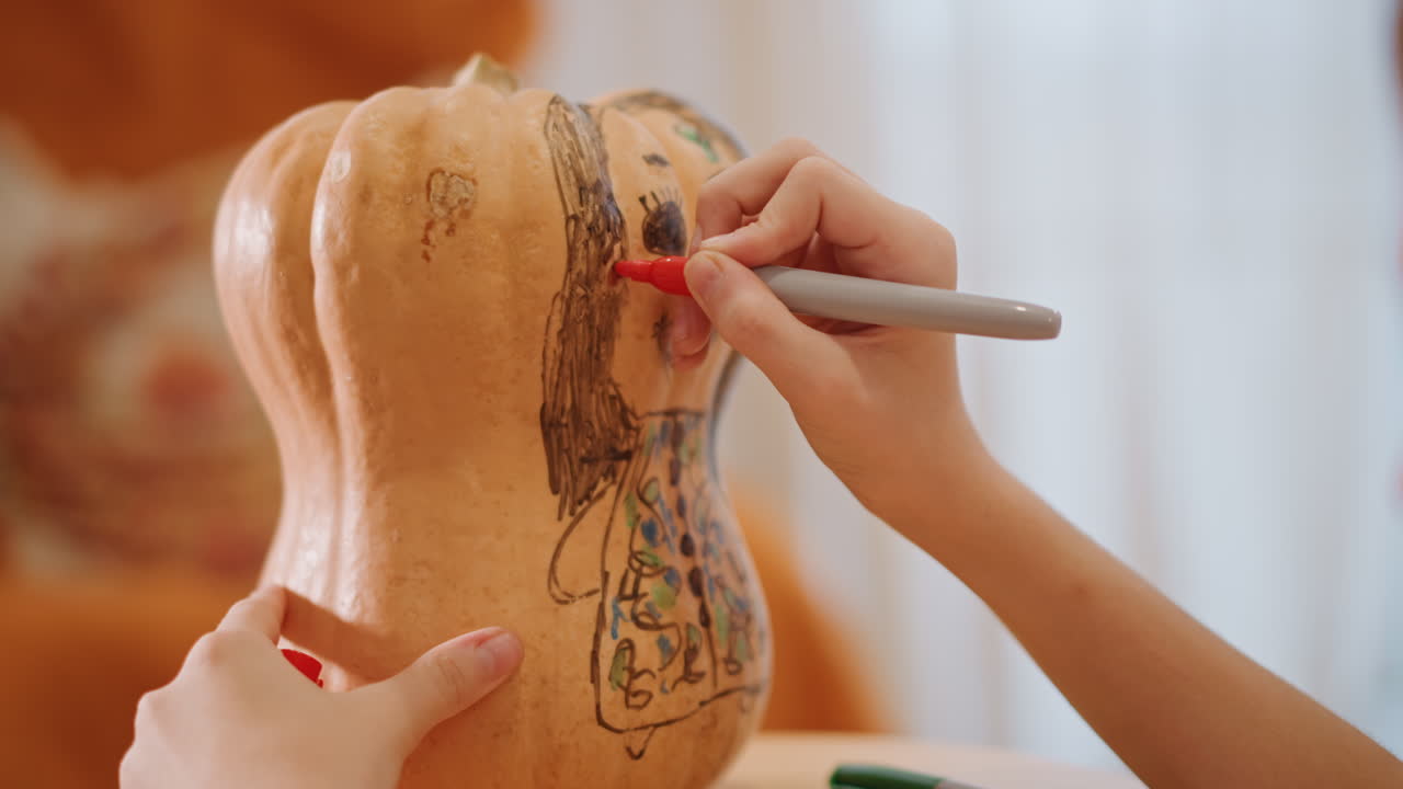 Manos dibujando una cara en una calabaza en un ambiente acogedor de cocina, esmalte de uñas rojo guiando el rotulador para esbozar cabello y sonrisa en una calabaza cacahuete, primer plano de una decoración creativa de bricolaje, luz otoñal cálida y desenfocada.