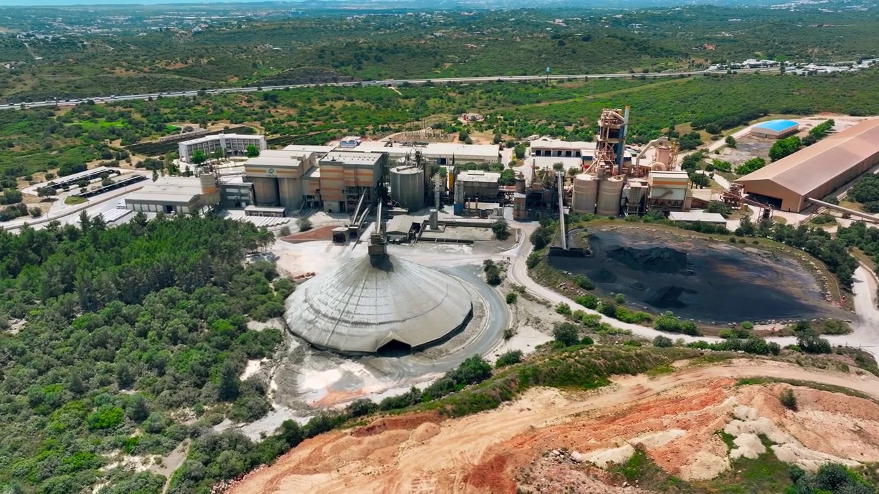 Aerial view of cement production facility showing limestone quarry operations and aggregate processing. Heavy machinery and conveyor systems transport materials across the site.