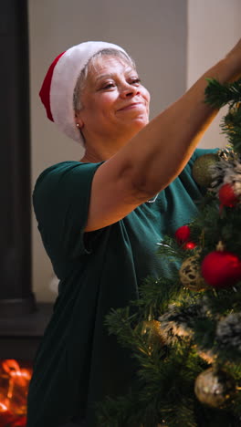 vertical video: smiling woman decorating christmas tree at home, wearing santa hat