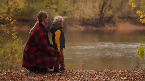 Descanso-Familiar-En-El-Bosque-De-Otoño,-La-Madre-Y-El-Hijo-Pequeño-Están-Viendo-El-Lago-Parado-En-La-Costa-Con-Pasto-Seco-Y-Follaje-De-árboles-Amarillentos-Alrededor