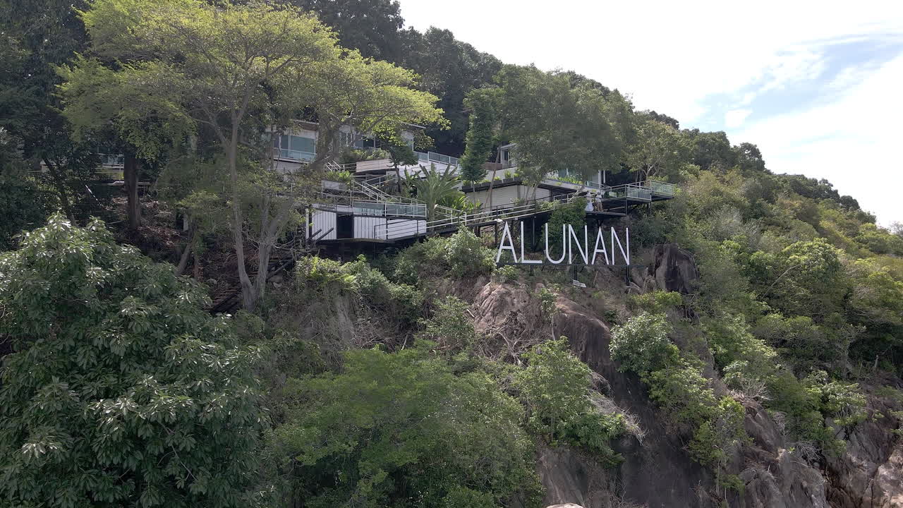 Slow aerial flight towards a large Alunan Resort sign above cliffs above a blue ocean. Rooms are hidden in the jungle behind the sign. Filmed on the remote island Pergentian Kecil, Malaysia.
