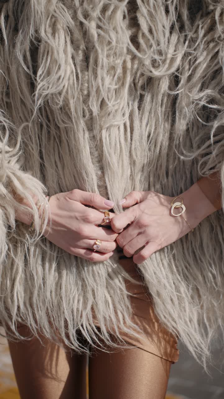 A woman gracefully slides her hands down a shaggy, textured fur coat, adorned with elegant gold jewelry, including rings and a bracelet, highlighting a chic and luxurious aesthetic - vertical, slowmo