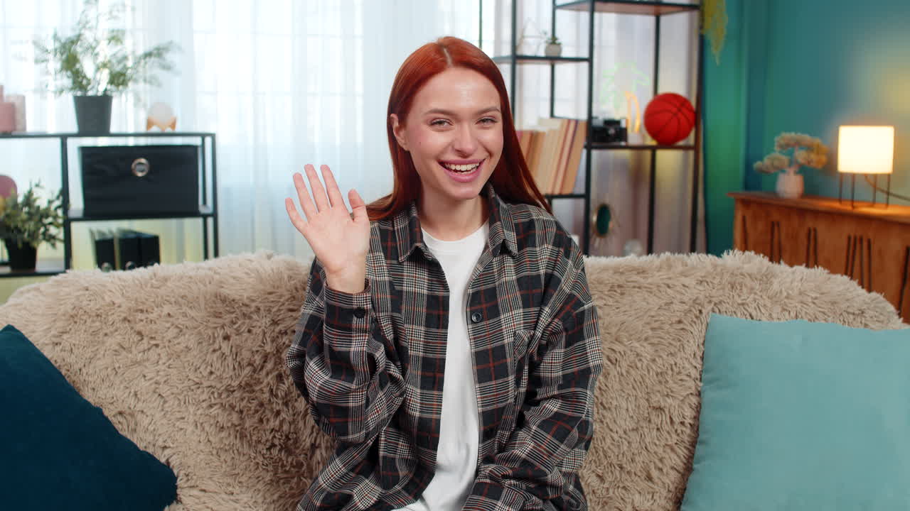 Woman sits on sofa waving in camera with cheerful smile greeting with joy during online connection