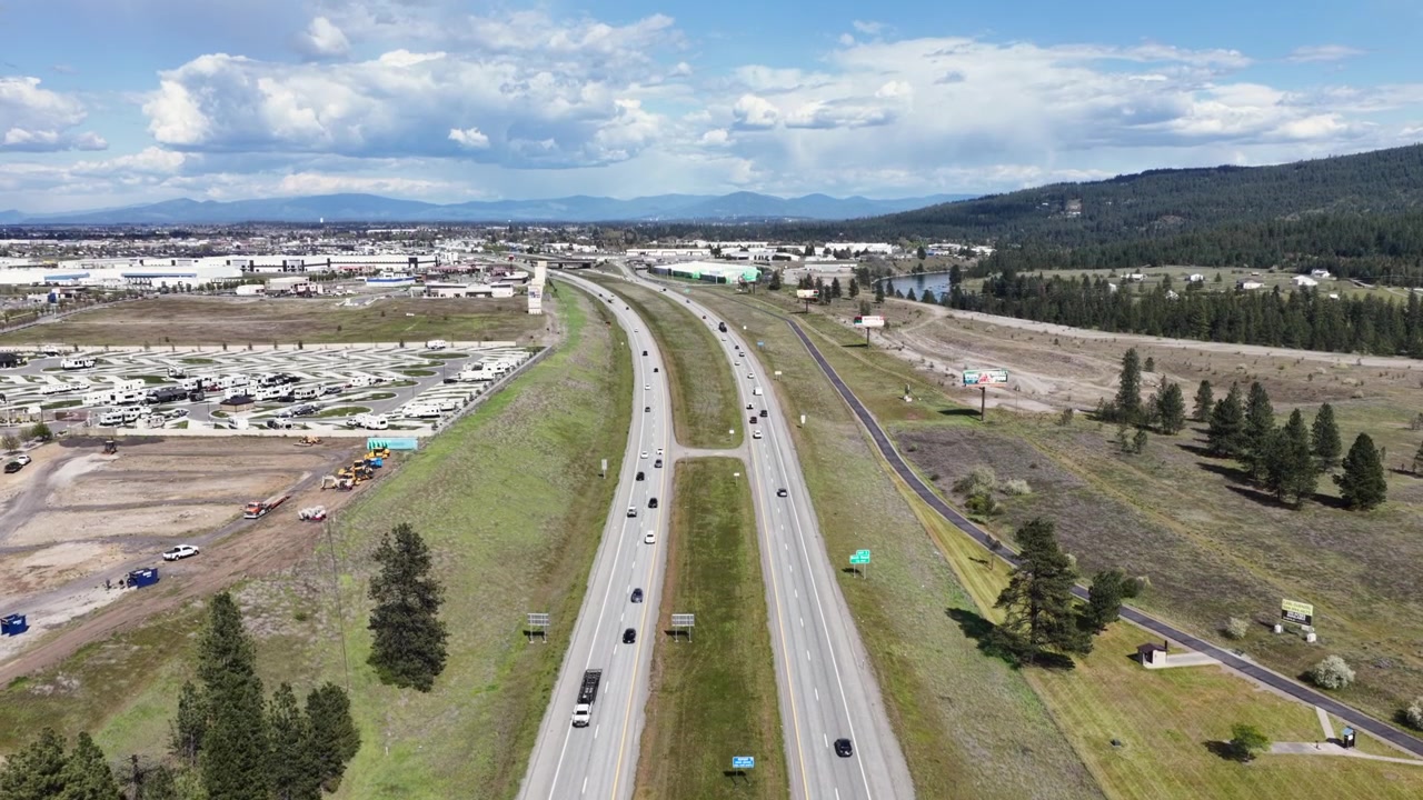 Deskripsi Rekaman Stok: Lalu lintas jalan raya yang sibuk di Interstate 90 mengalir melalui perbukitan bergelombang yang indah dan lanskap hijau di bawah langit cerah di Post Falls, Idaho, Amerika Serikat. Udara.