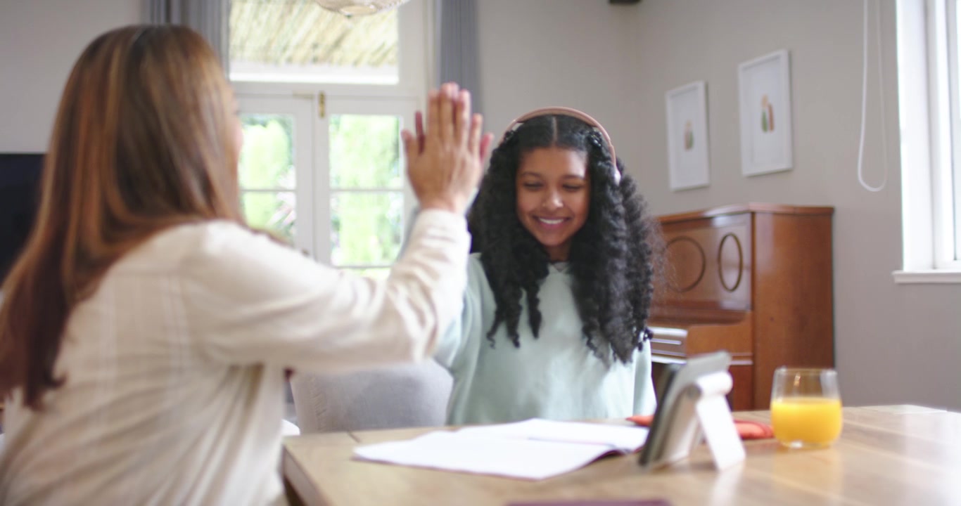 Madre afroamericana e figlia adolescente che si battono il cinque dopo un successo, studiando un quaderno di esercizi al tavolo