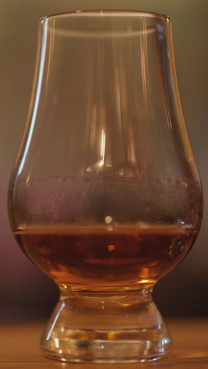 Un singolo bicchiere di whisky con liquido ambrato ruota lentamente su una superficie di legno, illuminato da una luce ambientale soffusa e calda e uno sfondo sfocato.