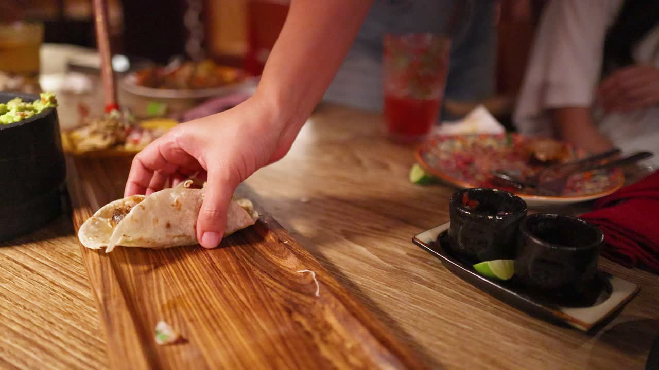 A hand reaches for a taco on a wooden table in a warmly lit Mexican restaurant, surrounded by dishes and drinks