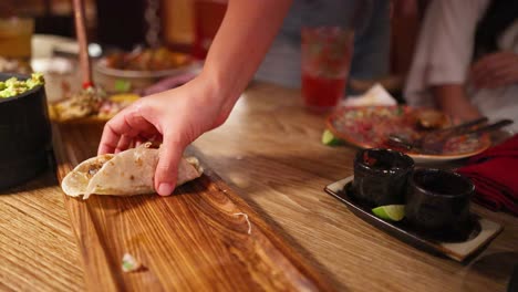 a hand reaches for a taco on a wooden table in a warmly lit mexican restaurant, surrounded by dishes and drinks