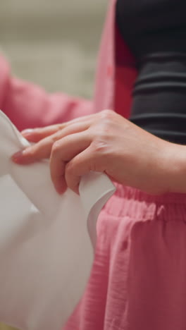 vista inferior del cuerpo de una dama con una camiseta rosa desabrochada evaluando cuidadosamente una camisa polo blanca, acercándola a ella para su inspección, el fondo muestra una vista borrosa de otras ropas