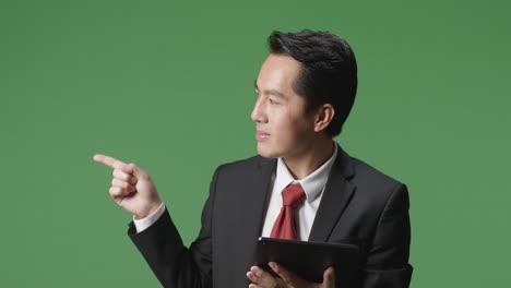 an asian business man using tablet and pointing to the side on green screen background in the studio
