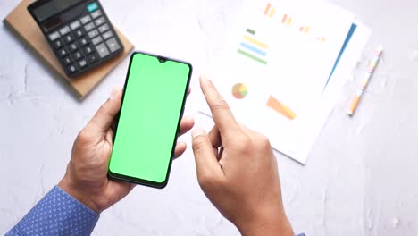 persona con un teléfono inteligente con pantalla verde, rodeada de documentos comerciales y una calculadora.