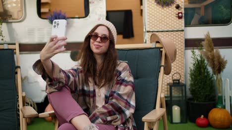 A-happy-girl-in-a-white-hat-and-plaid-shirt-sits-on-a-green-chair-against-the-backdrop-of-a-picnic-trailer-and-takes-a-selfie-using-a-white-smartphone-during-her-trip-outside-the-city-in-the-summer