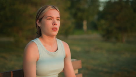 young tired lady sitting outdoors, looking down with exhaustion while blurred greenery fills background, golden sunlight reflects off her face