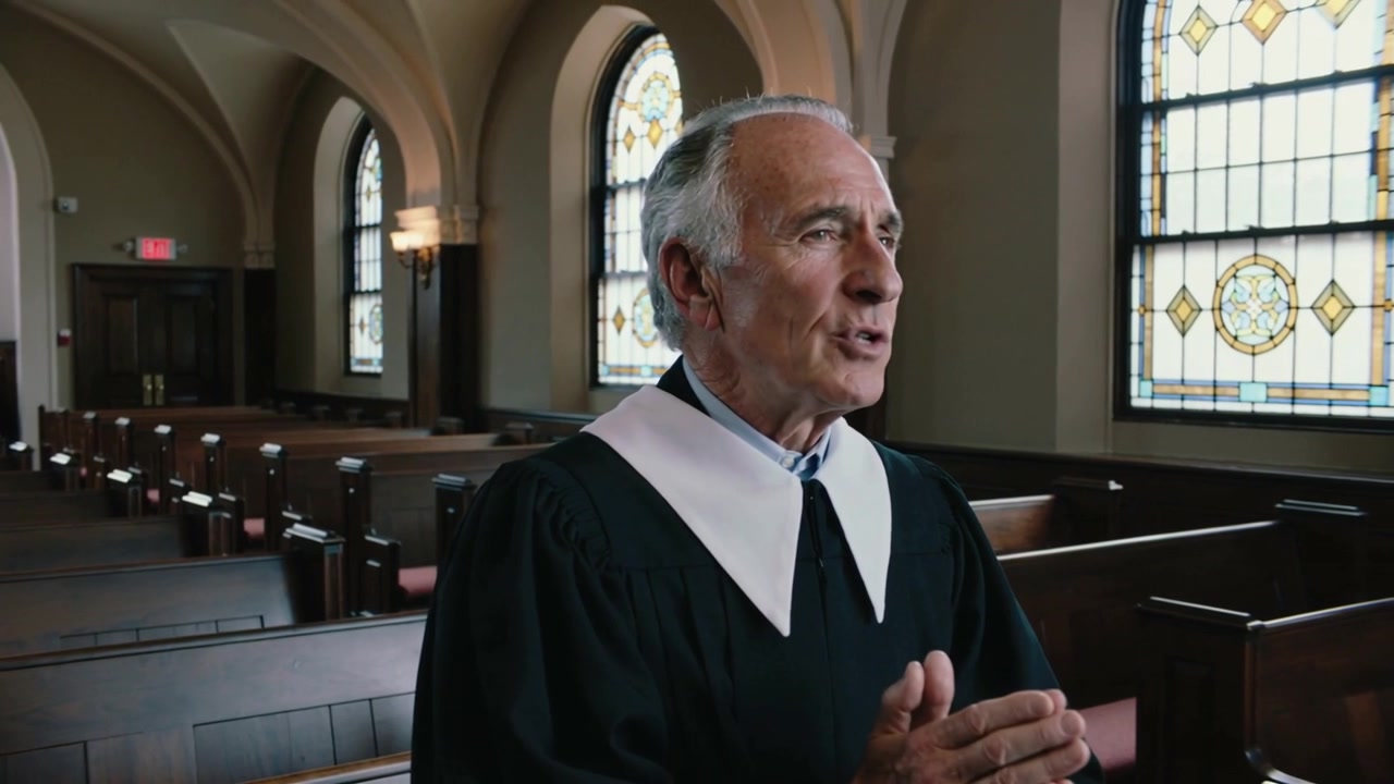 Ein älterer Mann in schwarzer Robe und weißem Kragen spricht und gestikuliert in einer Kirche, während das Licht durch Buntglas fällt.