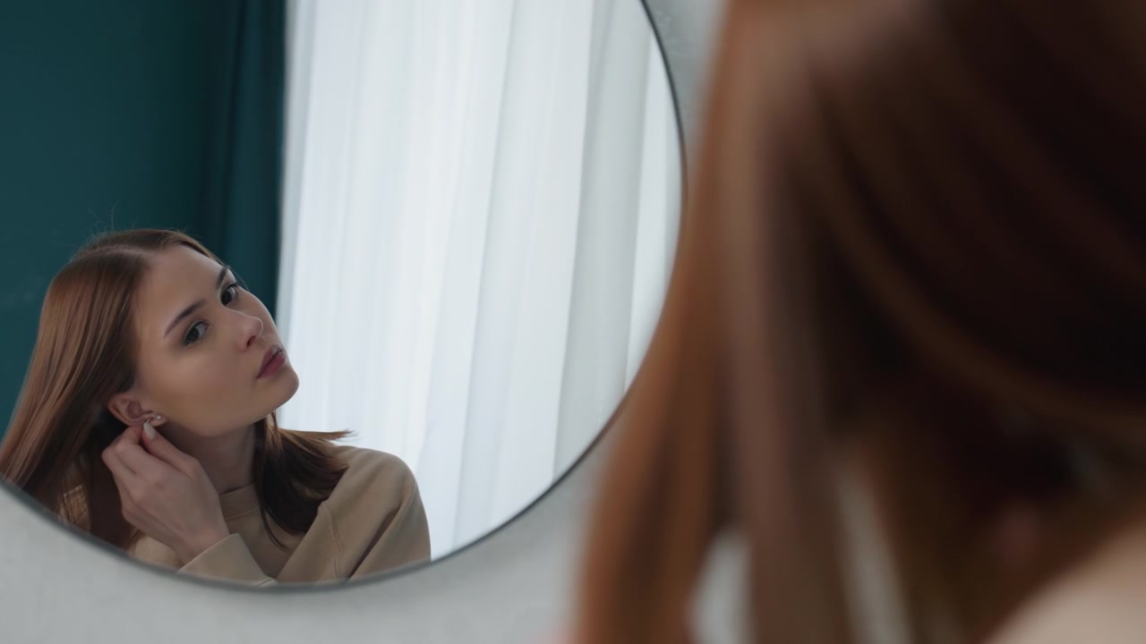 Giovane donna che ripassa la lezione allo specchio, pratica la postura e le espressioni facciali accanto alla luce soffusa della finestra e a una tenda color verde acqua, atmosfera da tutor concentrato con laptop e appunti impliciti, calma determinazione.
