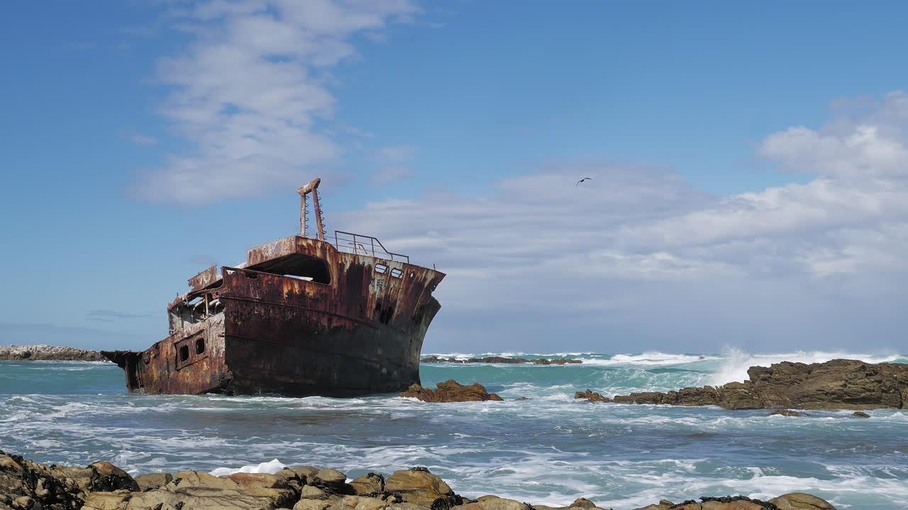 Old weathered shipwreck in shallows with waves running in, rocky coastline of L'Agulhas, South Africa, static shot