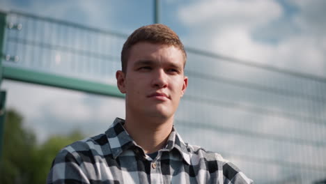 young man in plaid shirt looking around with focused expression, standing in outdoor sports area with blurred fence, greenery, and clear sky in background