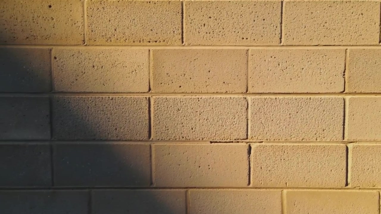 Close-up of a beige cinder block exterior wall lit from the right, showing rough porous surfaces, rectangular block pattern and horizontal mortar joints. Warm