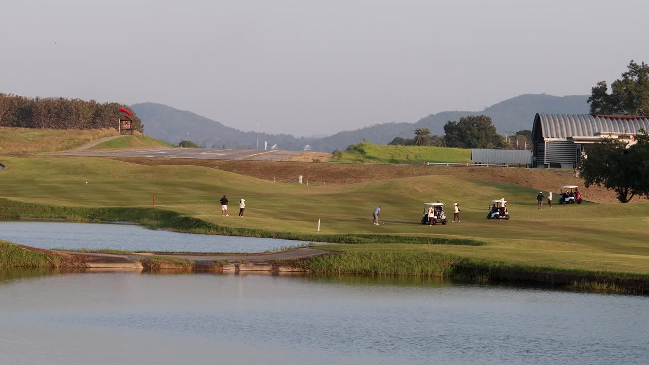 Golfers and carts on lush course near lake, mountains, and sunset, wide static shot