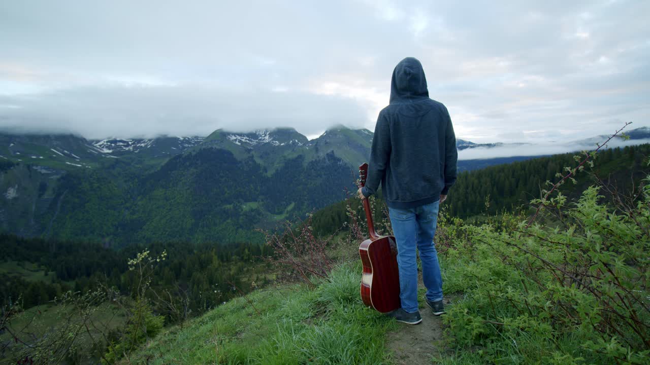 A man is standing at the top of a mountain with his guitar looking at the view