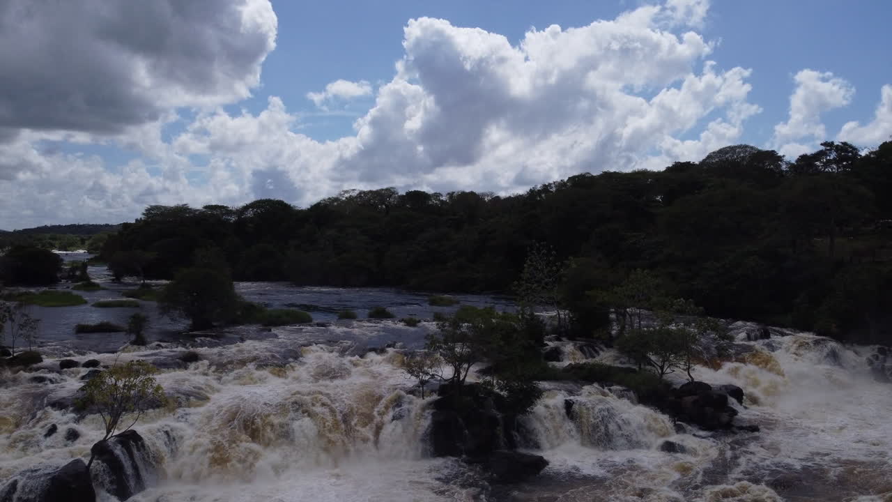 el fotograma aéreo captura fragmentos de árboles y rocas incrustados dentro de los rápidos del río mientras la cámara sube, revelando una perspectiva más amplia