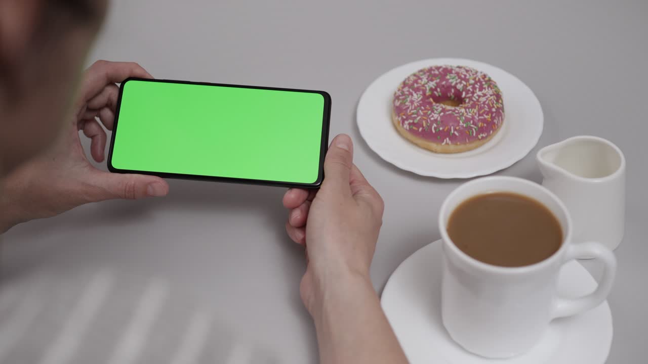mujer sentada en la mesa de café donut usando un teléfono inteligente con pantalla verde de clave de croma, desplazándose a través de las redes sociales medios de comunicación en línea tienda internet. teléfono inteligente en modo horizontal con maqueta de pantalla verde.