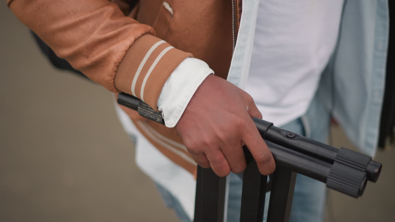 Closeup hand gripping tripod stand on sidewalk, casual jacket and jeans, DJ transporting equipment through urban street, candid motion and hustle, focus on carry and setup, energetic lifestyle vibe