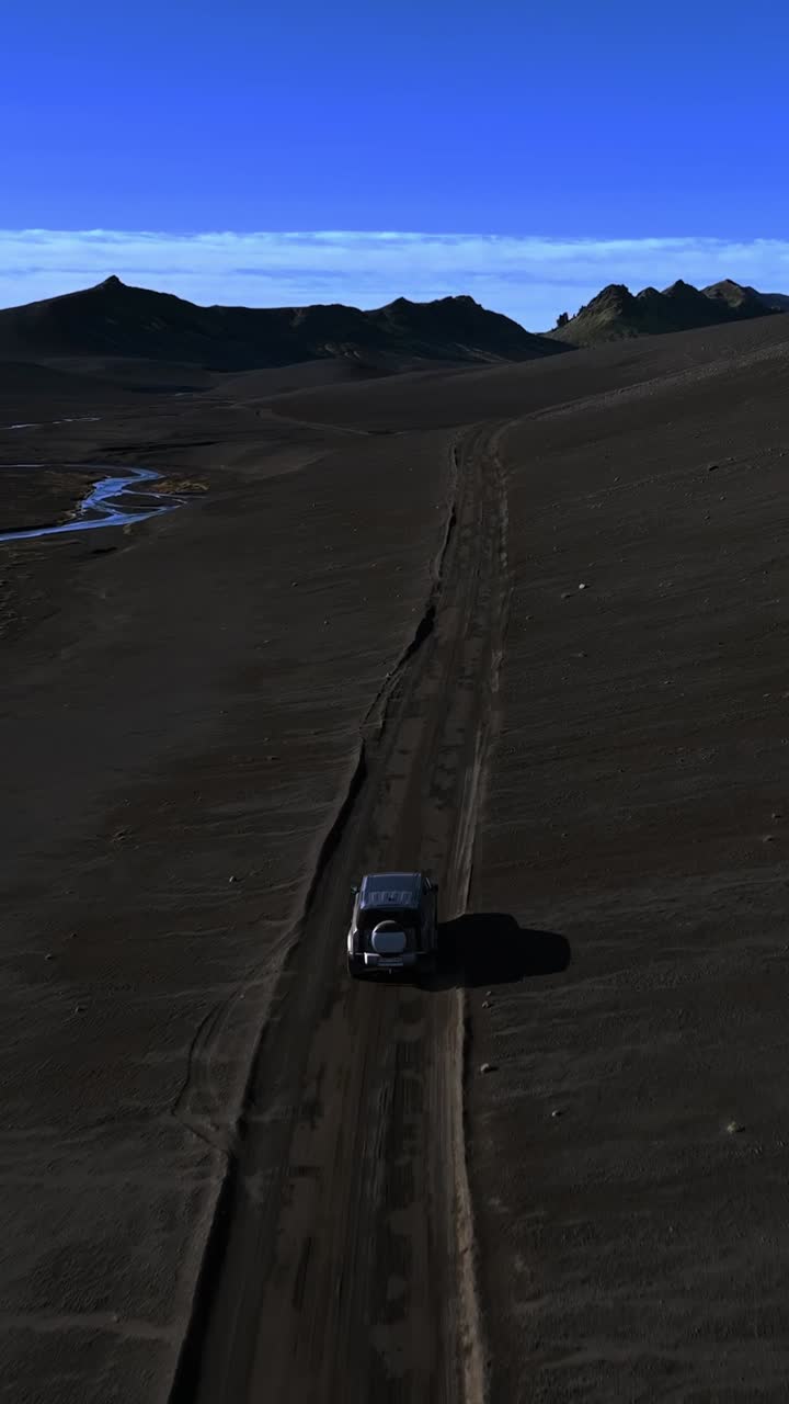 Vista aerea verticale da drone che segue un SUV 4x4 su una stretta strada sterrata tra le colline di sabbia nera islandesi, con un fiume tortuoso e vette vulcaniche in lontananza