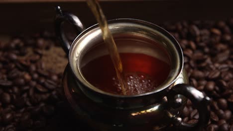 Luxurious-rich-coffee-pouring-into-ornate-silver-cup-on-dark-ground-aromatic-coffee-beans-close-up