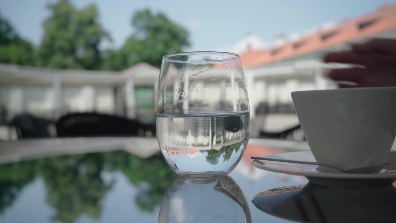 Bicchiere d'acqua e tazza di caffè su un tavolo riflettente di un caffè all'aperto mentre una mano si allunga in una calda giornata estiva