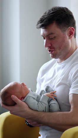 caring caucasian father waving his newborn son. man sits in chair dandling tiny kid in blue romper. vertical video