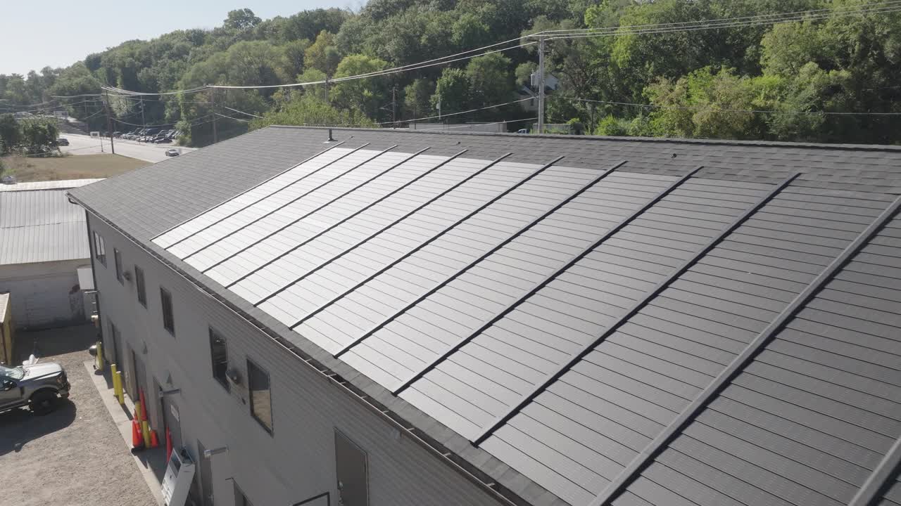 드론은 초록색과 산업 구조로 둘러싸인 큰 상업용 지붕에 설치된 태양 전지를 보여줍니다.