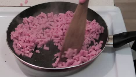 person cooking ham small pieces in a frying pan on electric stove, mixing with a wooden spatula, still shot 50fps