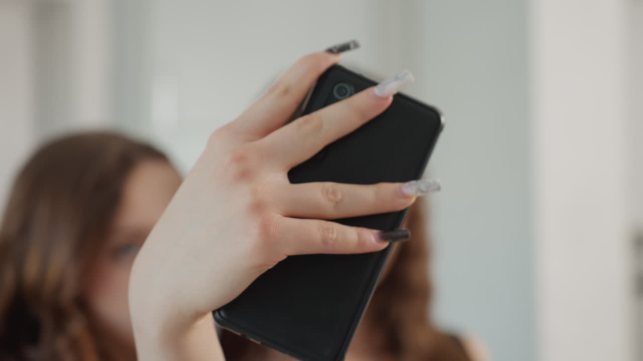 Hand Holding Phone With Prominent Acrylic Nails, Foreground Features Nails And Phone With Blurred Background Faces, Close Perspective Showing Detailed Nails And Phone With Indistinct Faces Behind