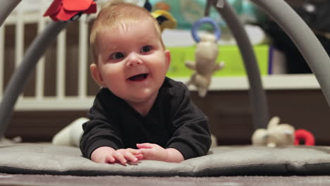 Happy-baby-girl-on-her-tummy-smiles-while-lying-under-hanging-toy-on-floor