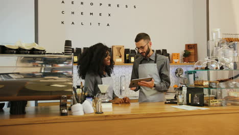 Junge-Kaukasische-Barista,-Die-Ihrem-Afroamerikanischen-Kollegen-Das-Neue-Menü-Des-Cafés-Auf-Einem-Tablet-Während-Des-Arbeitstages-Zeigt