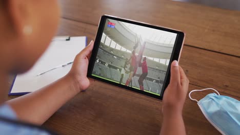 Composite-of-woman-sitting-at-table-with-face-mask,-watching-rugby-match-on-tablet