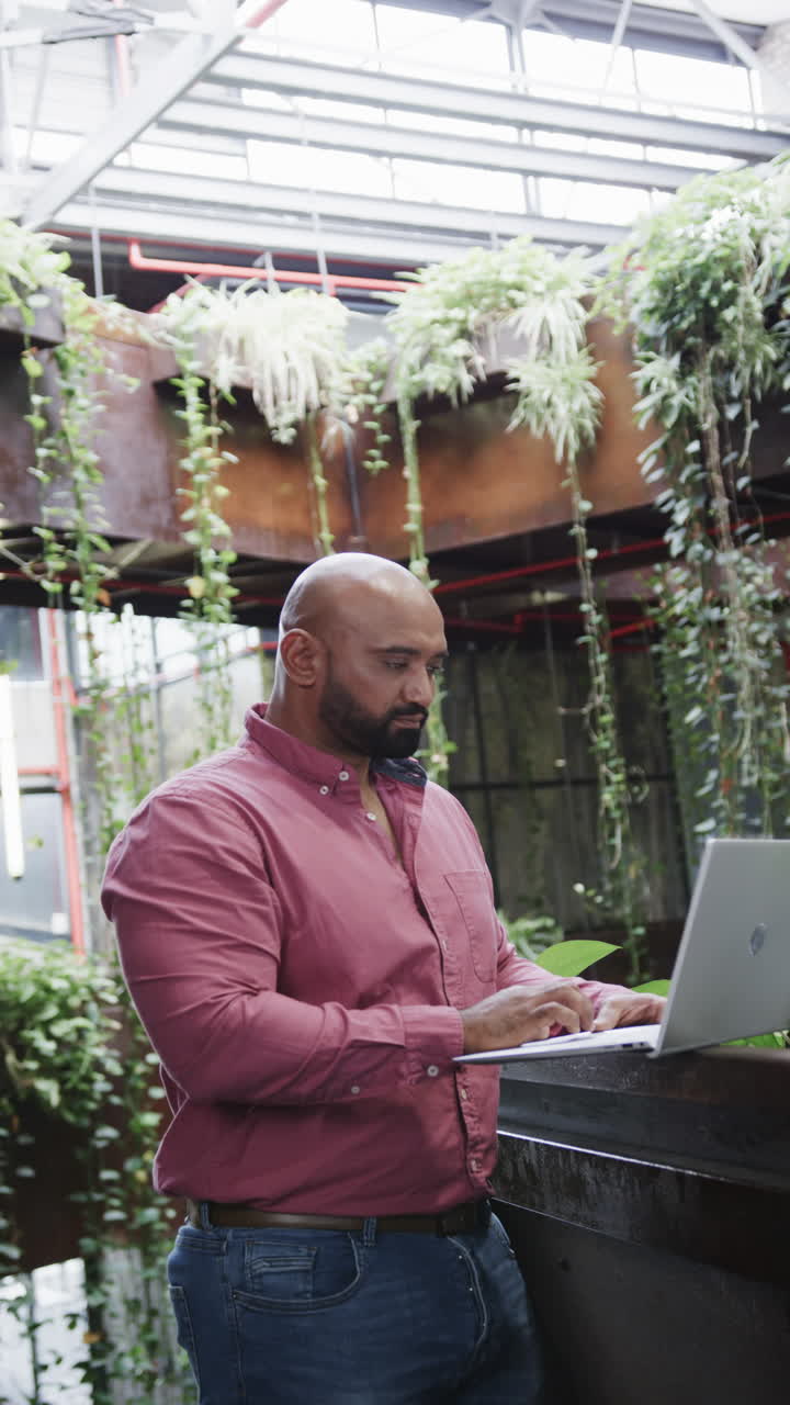 Vertical video of happy biracial male creative using laptop at casual office, in slow motion