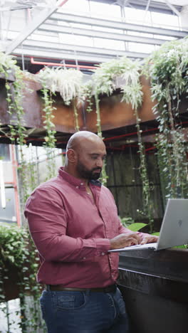 vertical video of happy biracial male creative using laptop at casual office, in slow motion